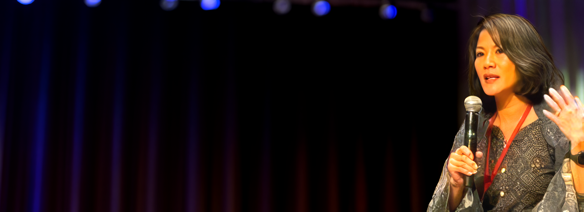 Ivy Chin speaking on stage at a keynote event, holding a microphone and gesturing to her audience against dramatic purple and blue stage lighting